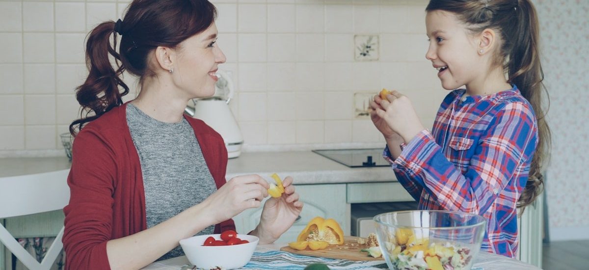 Happy family of young mother and cute cheerful daughter have fun grimacing silly with vegetables while cooking in the kitchen at home