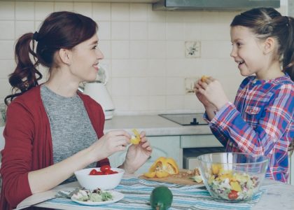 Happy family of young mother and cute cheerful daughter have fun grimacing silly with vegetables while cooking in the kitchen at home